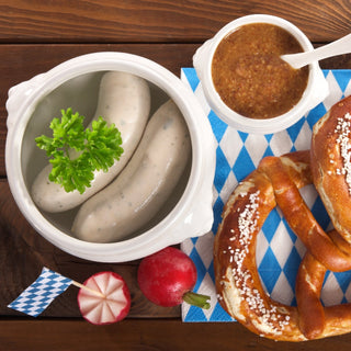 Sausages in a bowl with mustard, pretzels, and radishes on a wooden table.